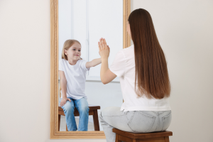 a woman is sitting on a stool looking at a mirror with her back to the camera. She is touching the mirror with her left hand. Reflected back at her is a small girl also touchingthe mirror. The image represents the course Healing The Inner Child