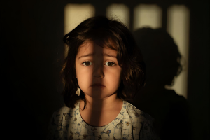 A young child looks dishevelled and sad, as they look longingly out of a window. The bands of light from the window emulate the bars of a cell.