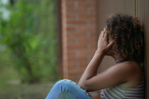 A woman sits with her back against a wall, with her head in her hands.